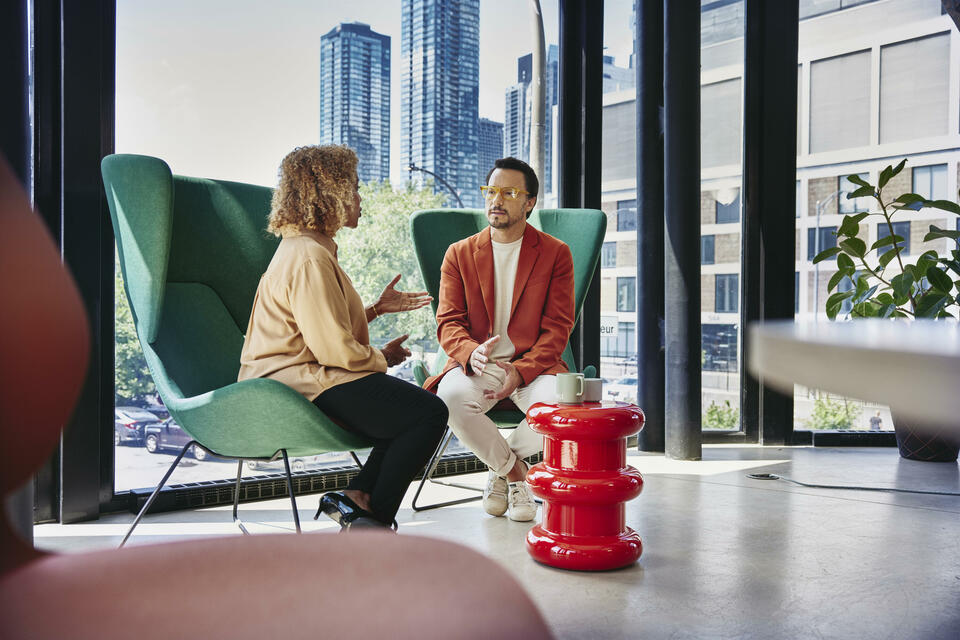 Male with glass and red blazer sitting in a green chair talking to a female sitting in a green chair
